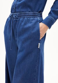 Blauwe denim broek met een elastische tailleband en trekkoord, voorzien van zijzakken en een klein merklabel aan de linkerkant.