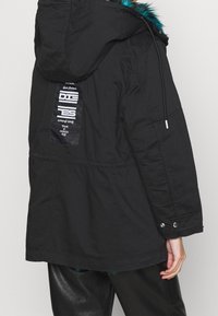 Svart parka med en huva med pälsinredning, elastisk midja och en logotyp på baksidan. Strukturerad tyg med tryckknappsdetaljer på ärmarna.