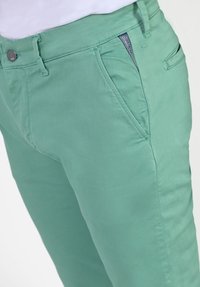 Lichtgroene katoenen broek met een fitted ontwerp, één voorzak en een klein merkje op de tailleband. Soepele textuur.