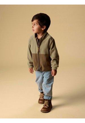 Mladi dječak gleda prema lijevo, nosi dvoslojnu smeđu jaknu na patent zatvarač, svijetloplave traperice i smeđe čizme, stoji na bež pozadini.