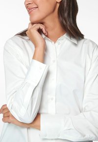 Weißes Hemd aus glattem Stoff, mit einem Button-Down-Kragen und langen Ärmeln. Sichtbare Knöpfe und klare Linien unterstreichen das Design.