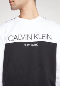 Felpa bianca e nera con design a girocollo. Presenta la scritta "CALVIN KLEIN" e "NEW YORK" stampata in caratteri in grassetto sul davanti.