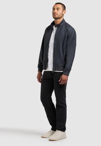 Férfi áll, tengerészkék cipzáras dzsekit, fehér pólót, fekete farmert és fehér tornacipőt visel, egyszínű világos háttér előtt.