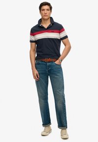 Polo navy con rayas rojas, blancas y grises, combinado con unos jeans azules deslavados. El modelo lleva un cinturón marrón y zapatillas ligeras color crema.