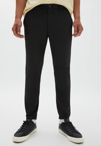 Homme portant un pantalon noir coupe slim, des baskets noires avec des semelles blanches et des chaussettes blanches, debout devant un fond uni.
