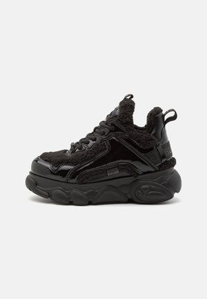 Black sneakers featuring a glossy patent leather upper with soft, textured fleece accents, thick sole, and round laces.