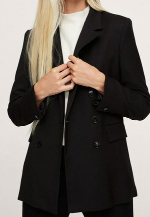 Personne blonde ajustant le devant d'un blazer noir à double boutonnage porté sur un haut blanc sur un fond uni.
