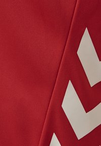 Roter Stoff mit einer vertikalen Naht und drei weißen, winkelförmigen Chevron-Symbolen, die entlang der Naht ausgerichtet sind.