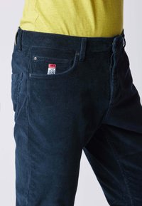 Pantaloni in corduroy blu navy con superficie testurizzata, dotati di tasche anteriori e un'etichetta sulla tasca posteriore, abbinati a una camicia gialla.