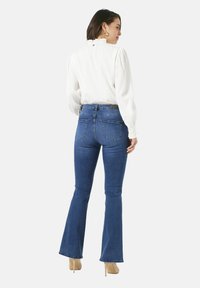 Vrouw in een witte blouse met lange mouwen, blauwe high-waisted flared jeans en beige hoge hakken, staande met haar rug naar de kijker gekeerd.