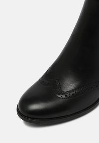 Bottine en cuir noir avec des détails de brogue et un bout rond sur fond blanc.