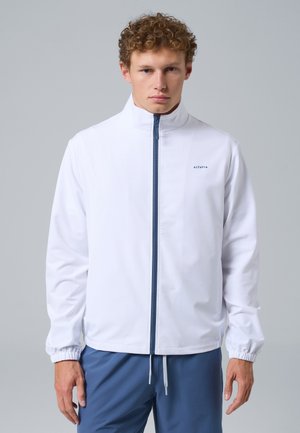 Giovane uomo con capelli ricci che indossa una giacca bianca con zip e pantaloni blu, in piedi davanti a uno sfondo grigio chiaro uniforme.