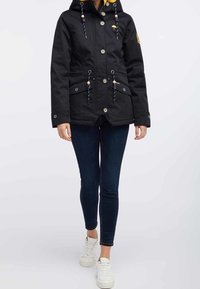 Czarna kurtka z kapturem zapinana na guziki z przodu, z troczkami i dwoma dolnymi kieszeniami. Posiada żółte podszycie oraz ozdobne elementy metalowe.