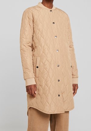 Homme portant un manteau matelassé beige mi-longueur avec boutons-pression et poignets côtelés, associé à un pantalon en velours côtelé beige.