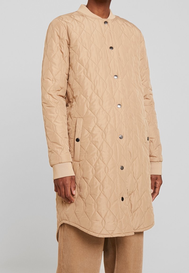 Homme portant un manteau matelassé beige mi-longueur avec boutons-pression et poignets côtelés, associé à un pantalon en velours côtelé beige.