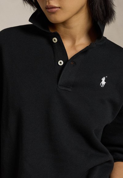 Nő fekete galléros pólót visel, melyen fehér hímzett póló játékos logó van a mellkasán, felállított gallérral és két begombolt gombbal.