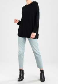 Schwarzer, strukturierter, übergroßer Pullover mit langen Ärmeln, kombiniert mit hellblauen, knöchellangen Jeans und schwarzen, hochhackigen Stiefeletten.