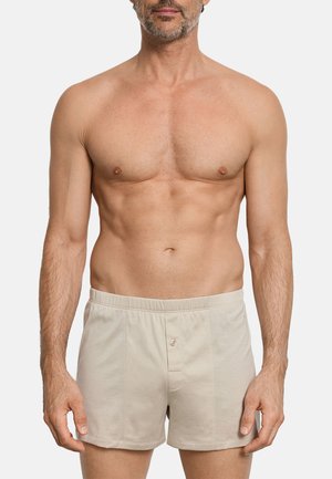 Uomo senza maglietta che indossa boxer beige con patta a bottoni, in piedi su uno sfondo bianco uniforme.