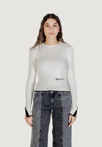 Top blanco de manga larga con canalé, escote redondo y puños de acento negros. Presenta texto de logo impreso en la parte delantera. Combinado con jeans de pierna ancha grises.
