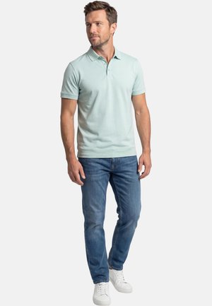 Mies, joka pukeutunut vaaleanvihreään lyhythihaiseen poolopaitaan, sinisiin farkkuihin ja valkoisiin lenkkareihin, seisoo rennosti käsien ollessa rentoina ja katsoo oikealle.