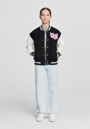 Giacca varsity nera e crema con bottoni a scatto, con la scritta "USA" in rosa sul petto; abbinata a jeans azzurri decorati e sneakers bianche.