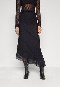 Vestido de encaje negro con mangas largas, diseño en capas, bordes festoneados y una textura transparente, combinado con botas de estilo cowboy negras.