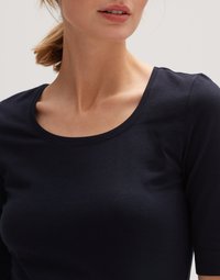 Vrouw met een lichte huid en haar naar achteren gebonden, die een nauwsluitend donkerblauw shirt met een ronde hals draagt, getoond van nek tot midden van de torso tegen een effen achtergrond.