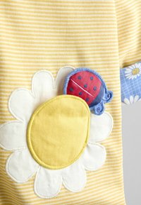 Geltonos ir baltos spalvos dryžuotas audinys su dideliu baltos ir geltonos gėlės aplikacija bei raudonos ir mėlynos spalvos boružės lopu ant mėlynos rankogalių su ramunėlių raštu.