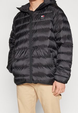 Man draagt een zwarte doorgestikte pufferjas met een ritssluiting aan de voorkant en beige broek, staand met handen in de zakken.