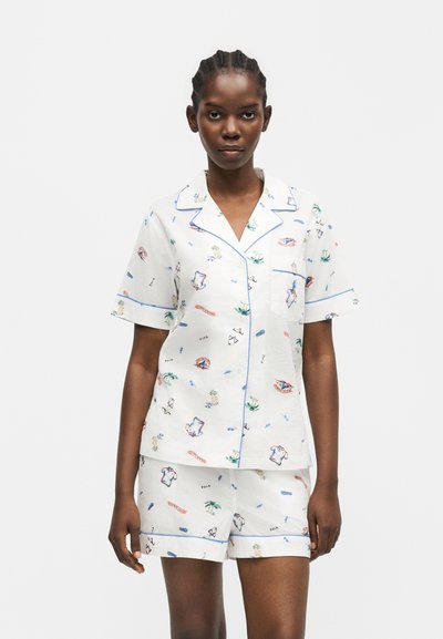 Woman wearing white short-sleeve pajama set with blue piping and colorful small prints, standing against a plain light background.