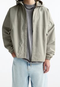 Veste à capuche gris clair en tissu lisse et résistant à l'eau, avec des poignets élastiques, des poches latérales et une coupe décontractée sur un t-shirt gris.