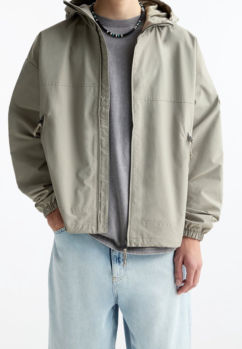 Veste à capuche gris clair en tissu lisse et résistant à l'eau, avec des poignets élastiques, des poches latérales et une coupe décontractée sur un t-shirt gris.