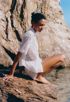 Jonge vrouw in witte eyelet outfit die blootsvoets op een rots zit bij helder water met een grote met zon beschenen rots achter haar.