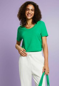 Camiseta de algodón verde de mangas cortas y cuello redondo, combinada con pantalones blancos con cordón. El fondo es de un tono violeta claro.