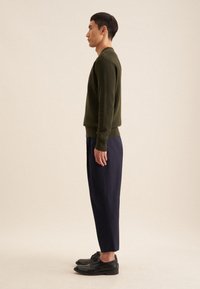 Pull en tricot vert olive avec des poignets côtelés, associé à un pantalon cropped bleu marine. Le mannequin porte des chaussures à lacets noires. Vue de côté, fond neutre.