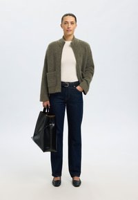 Ženska stoji, oblečena v olivno zeleno jopico, belo majico, temno modre kavbojke, črni pas, črne čevlje in drži veliko črno usnjeno torbo za na rame.