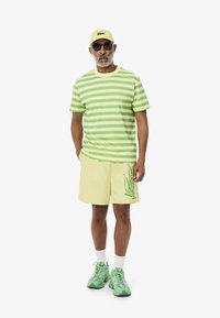 Gestreepte groene en gele t-shirt, gele shorts met grafische details, groene sneakers en een gele pet met logo. Gladde stof en casual pasvorm.