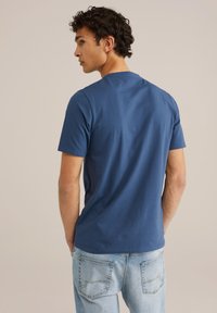 Moška kratkosleeve majica v temno modri barvi iz bombaža z okroglim izrezom, gladko teksturo in sproščenim krojem, v kombinaciji s svetlo modrimi kavbojkami.