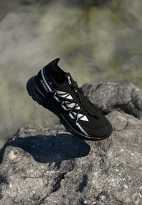 Black athletic shoe with a textured upper, reflective white accents, and a padded collar, floating above a rocky surface.