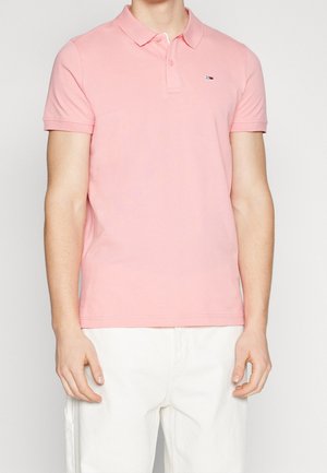 Homme portant un polo rose clair à manches courtes avec col et un pantalon blanc, debout devant un fond clair uni.