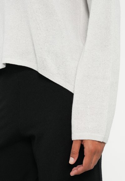 Pull en maille gris clair avec une texture côtelée, présentant un ourlet asymétrique. Pantalons en tissu noir visibles en bas, montrant une texture lisse.