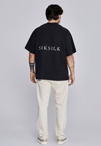 Črna prevelika majica s logotipom "SIKSILK" na hrbtni strani, kombinirana s kremasto obarvanimi trenirkami in čevlji z sivimi poudarki. Mehka tkanina.