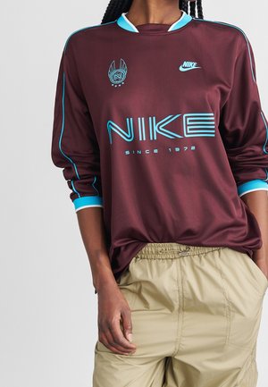 Osoba nosząca bordową, długorękawową koszulkę Nike z niebieskimi akcentami i napisem „NIKE since 1972”, zestawioną z beżowymi spodniami.