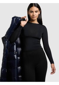 Sieviete ar gariem tumšiem matiem, ģērbta melnā cieši piegulošā garroku kreklā un melnās biksēs, pārlikusi tumši zilu pūkainu jaku pār plecu.