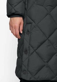 Left hand beside dark gray quilted jacket with large diamond stitching and black snap buttons on a white background.