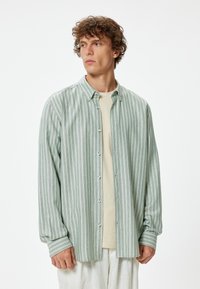 Camisa de botones a rayas verdes y blancas con un corte relajado, de mangas largas y puños. Llevada sobre una camiseta de color claro. Textura de tejido suave.