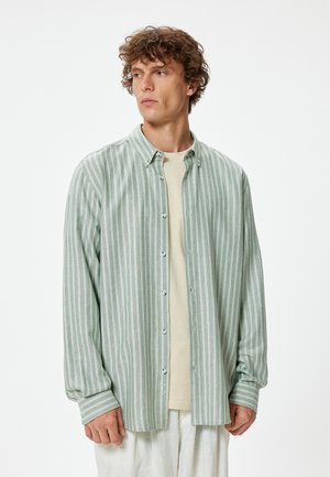 Camisa de botones a rayas verdes y blancas con un corte relajado, de mangas largas y puños. Llevada sobre una camiseta de color claro. Textura de tejido suave.