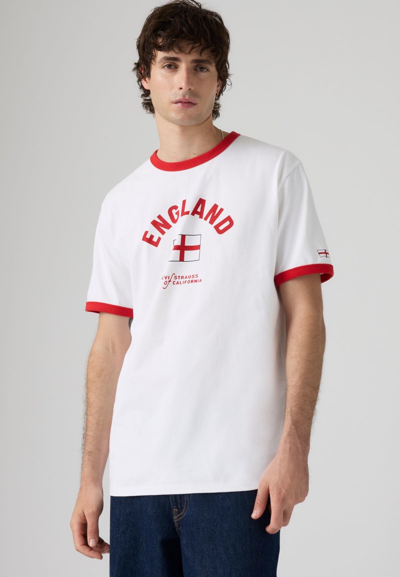 Νέος άνδρας που φοράει λευκό T-shirt με κόκκινες λεπτομέρειες, κείμενο "ENGLAND" και γραφικό της σημαίας της Αγγλίας, σε συνδυασμό με σκούρο μπλε τζιν σε απλό φόντο.