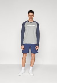 Γκρίζο μακρύ μανίκι μπλούζα με ναυτικές ράγες, με το λογότυπο "HUMMEL"; σε συνδυασμό με ναυτικά σορτς και λευκά αθλητικά παπούτσια.