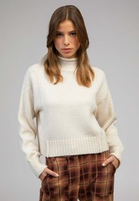 Pull en tricot blanc avec un col montant, des poignets et un ourlet côtelés. Associé avec un pantalon à carreaux marron présentant un motif à carreaux.